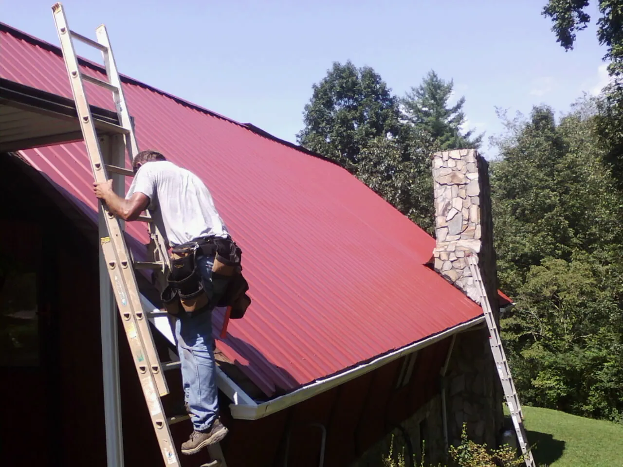 Roofer on a ladder beside a red metal roof and stone chimney — Soffit & Fascia Repair by Roofing Brothers in Tavares